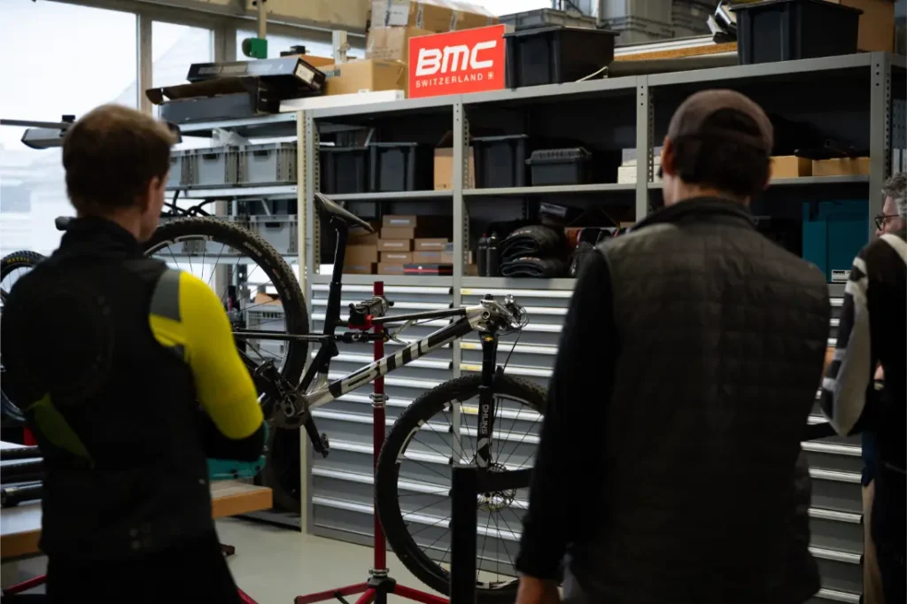 Dos personas se encuentran junto a una bicicleta colocada en un soporte de reparación, en un taller repleto de estanterías, herramientas y un letrero rojo de BMC Switzerland: un entorno en el que la revolución de la industria ciclista cobra vida con innovadoras ruedas de 32”.